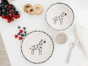 Kinder-Geschirrset aus weißer Keramik mit schwarzem Zebra-Motiv und grafischem Rand, bestehend aus Teller, Schale und Tasse, in der Draufsicht mit Beeren und Löffel arrangiert