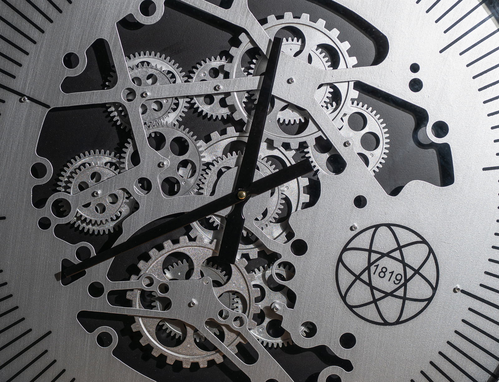 Nahaufnahme einer silberfarbenen Wanduhr aus Metall mit sichtbaren Zahnrädern und schwarzem Zifferblatt, frontal betrachtet.