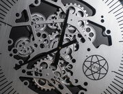 Nahaufnahme einer silberfarbenen Wanduhr aus Metall mit sichtbaren Zahnrädern und schwarzem Zifferblatt, frontal betrachtet.