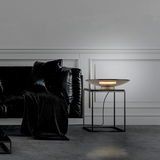 Moderne Tischleuchte PURE Moon auf einem quadratischen Beistelltisch, seitliche Perspektive, in einem stilvollen Wohnzimmer mit schwarzem Sofa und eleganter Wandverkleidung.
