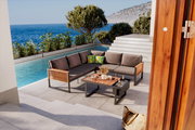 Lounge-Set auf einer Terrasse mit Meerblick, bestehend aus einem Ecksofa mit grauen Kissen und einem Holztisch, umgeben von Pflanzen und einem Pool. Perspektive von der Seite.