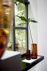 Zwei braune Glasvasen auf einer Fensterbank, eine mit einer langen grünen Pflanze, seitliche Perspektive