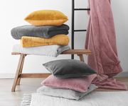 Verschiedene Felldecken und Kissenhüllen in Gelb, Grau, Weiß und Rosa auf einer Holzbank und einem Teppich, mit einer Leiter im Hintergrund. Perspektive von vorne.