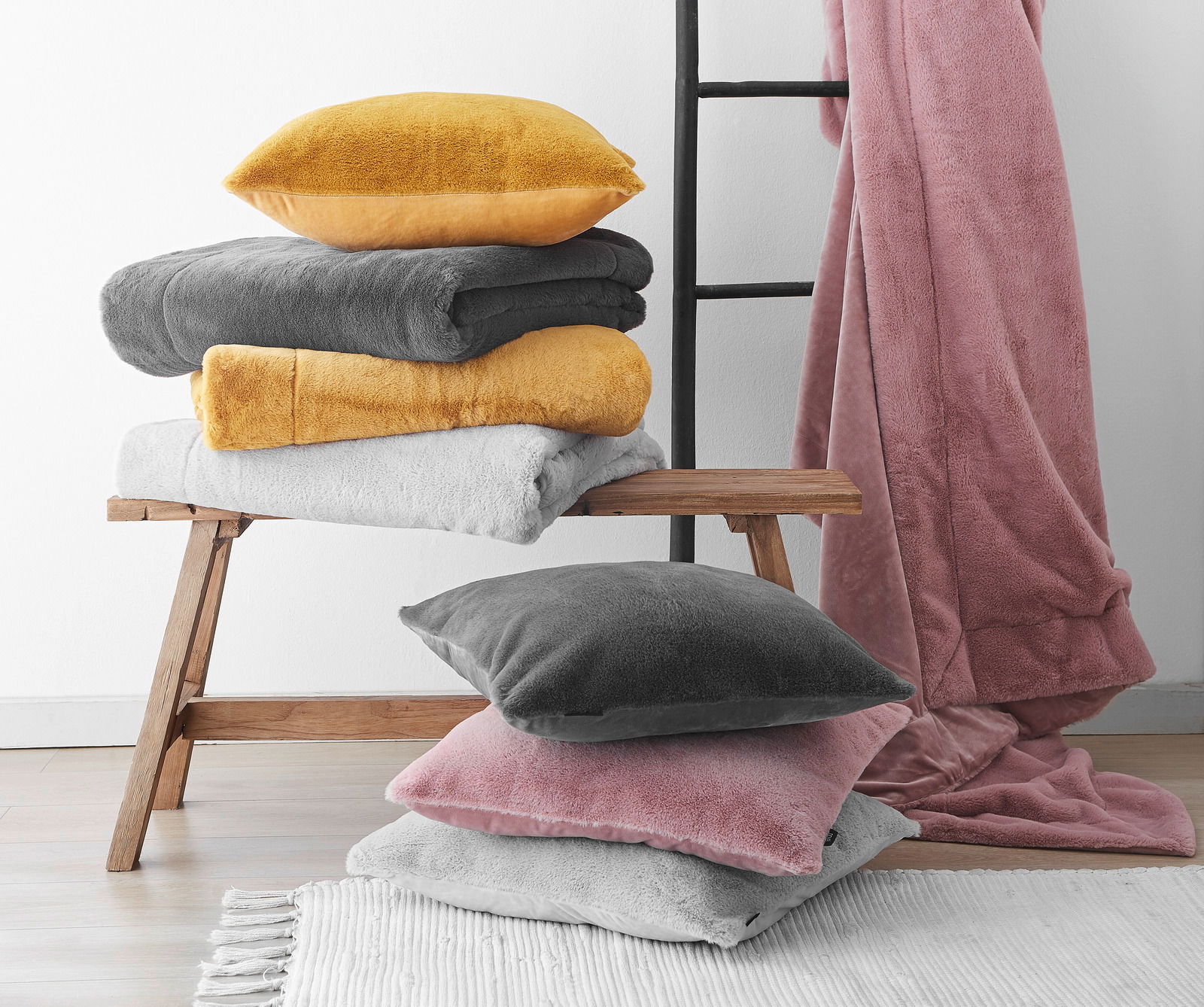 Verschiedene Felldecken und Kissenhüllen in Gelb, Grau, Weiß und Rosa auf einer Holzbank und einem Teppich, mit einer Leiter im Hintergrund. Perspektive von vorne.