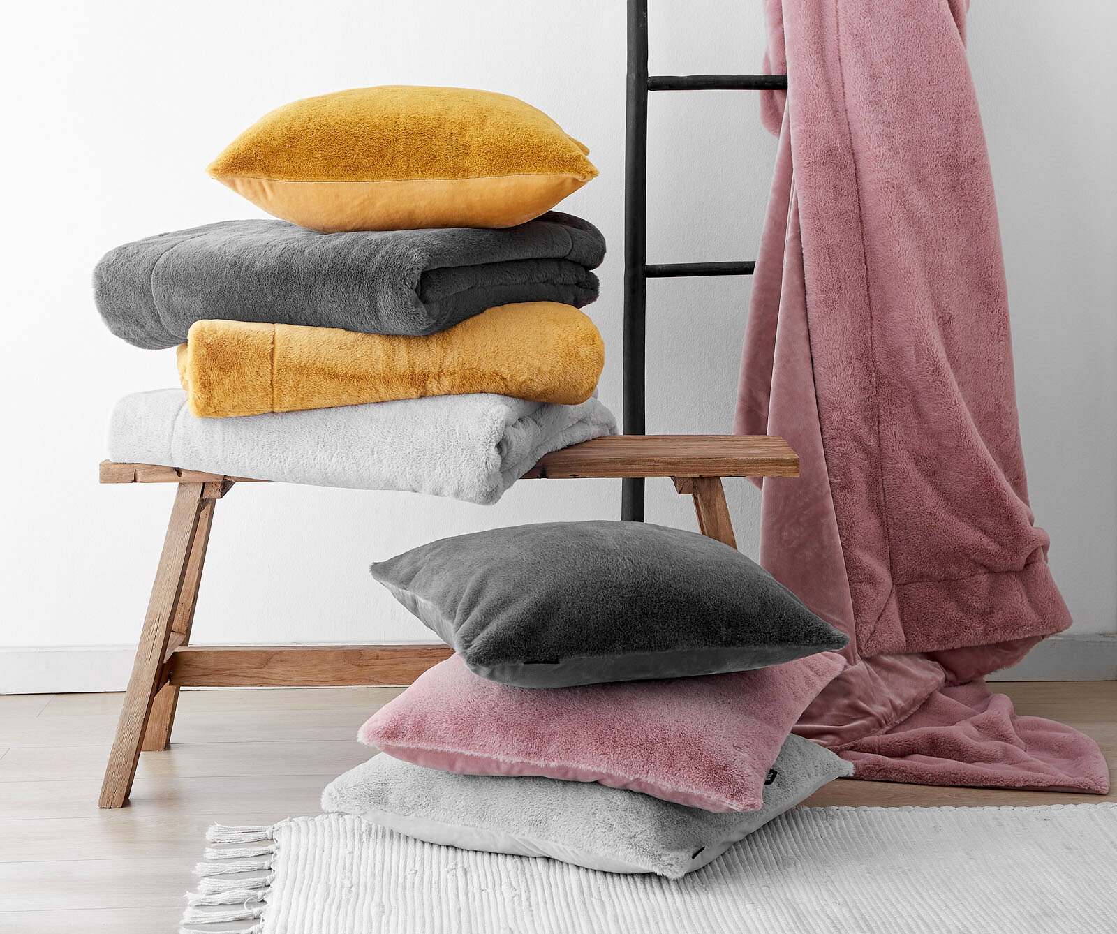 Verschiedene Felldecken und Kissenhüllen in Gelb, Grau, Weiß und Rosa auf einer Holzbank und einem Teppich, mit einer Leiter im Hintergrund. Perspektive von vorne.