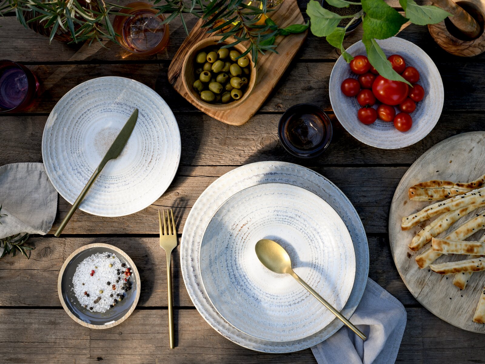 Draufsicht auf rustikales Outdoor-Tischgedeck mit weißen Keramiktellern, goldfarbenem Besteck, Schalen mit Oliven und Cherrytomaten sowie Brot auf Holzplatte.