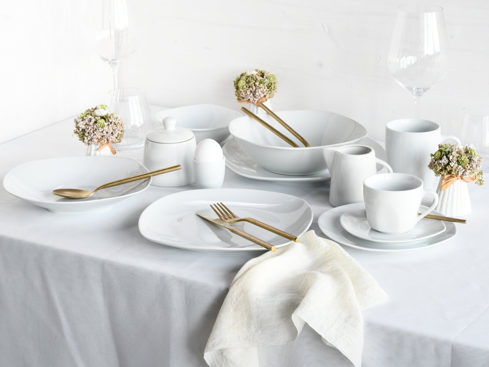 Elegant gedeckter Tisch mit weißem Porzellan-Geschirrset (Teller, Schalen, Tassen), goldfarbenem Besteck, Gläsern und weißen Servietten, aufgenommen aus leicht erhöhter Frontperspektive (3/4-Ansicht).