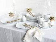 Elegant gedeckter Tisch mit weißem Porzellan-Geschirrset (Teller, Schalen, Tassen), goldfarbenem Besteck, Gläsern und weißen Servietten, aufgenommen aus leicht erhöhter Frontperspektive (3/4-Ansicht).