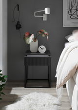 Moderne Nachtkommode in dunklem Design mit geriffelter Front, dekoriert mit einer Vase und einer Uhr, seitliche Perspektive.