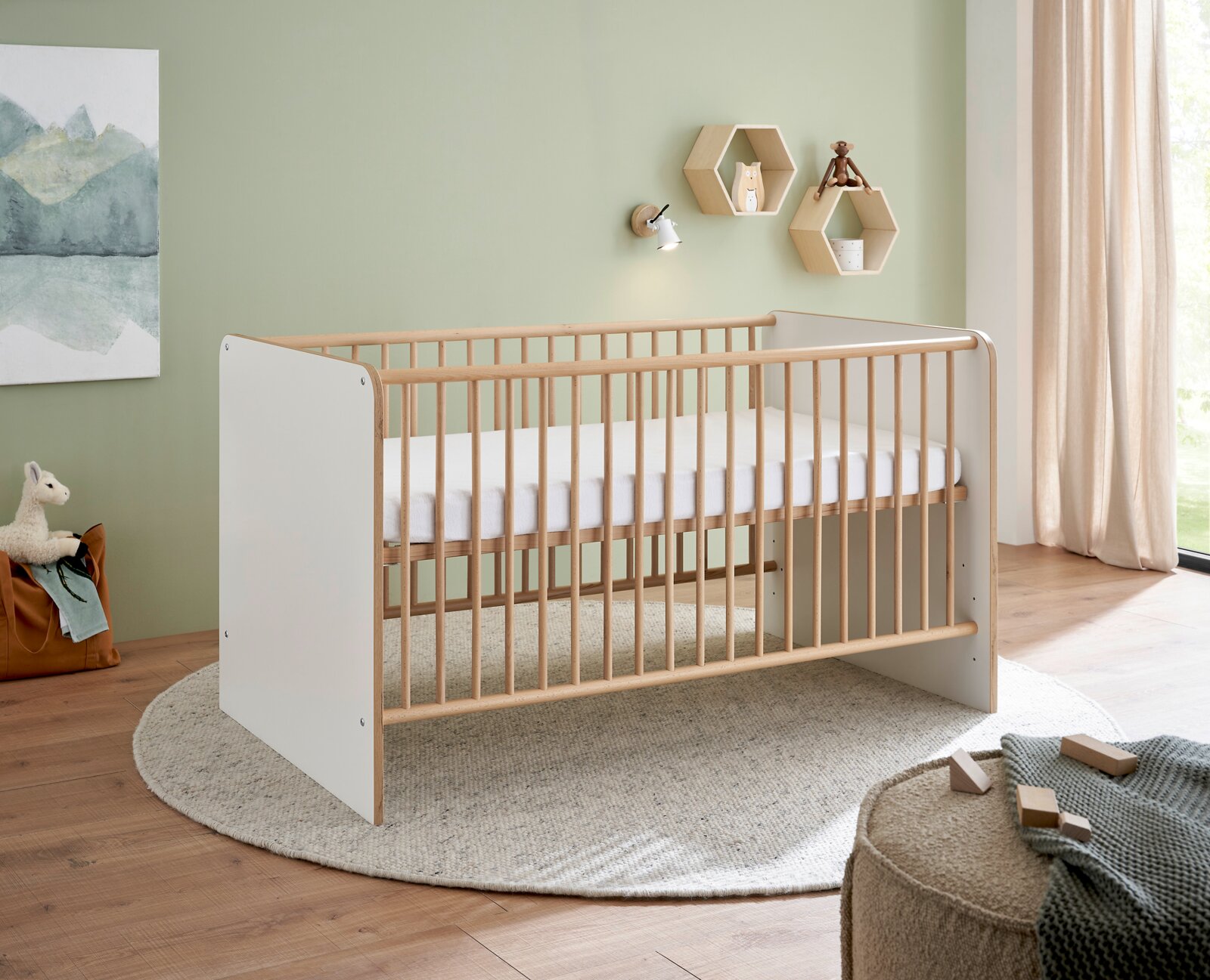 Modernes Babybett aus Holz mit weißen Seitenteilen und Naturholz-Gitterstäben im Kinderzimmer, schräg von vorne links fotografiert.