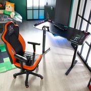 Gaming-Stuhl in Orange und Schwarz, seitliche Perspektive, neben einem Gaming-Tisch mit Tastatur und Controller.