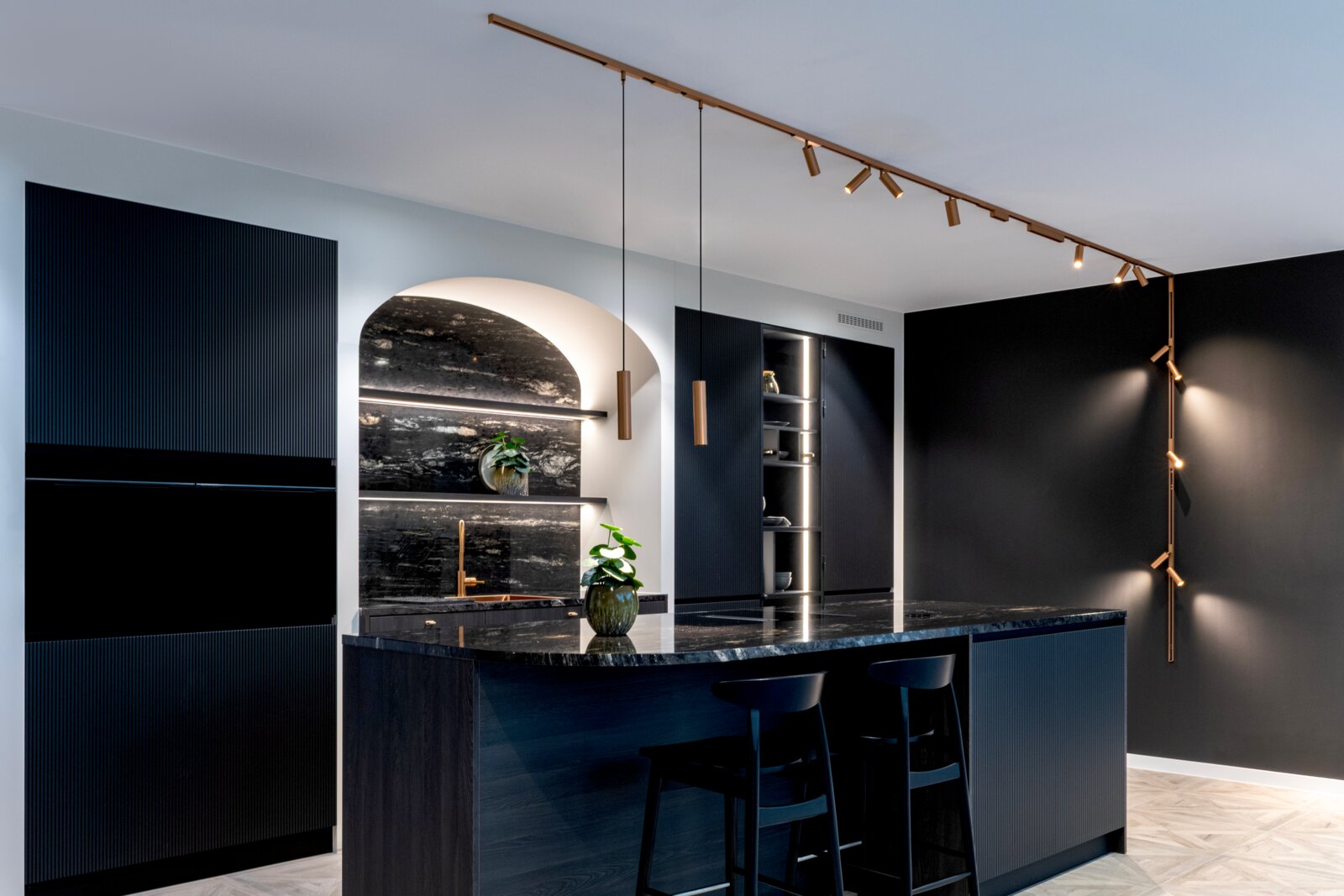 Moderne, dunkelblaue Designküche mit Kücheninsel und schwarzer Stein-Arbeitsplatte, goldfarbener Schienen- und Pendelbeleuchtung sowie Nischenregalen; Perspektive: leicht schräg von vorne aus Augenhöhe