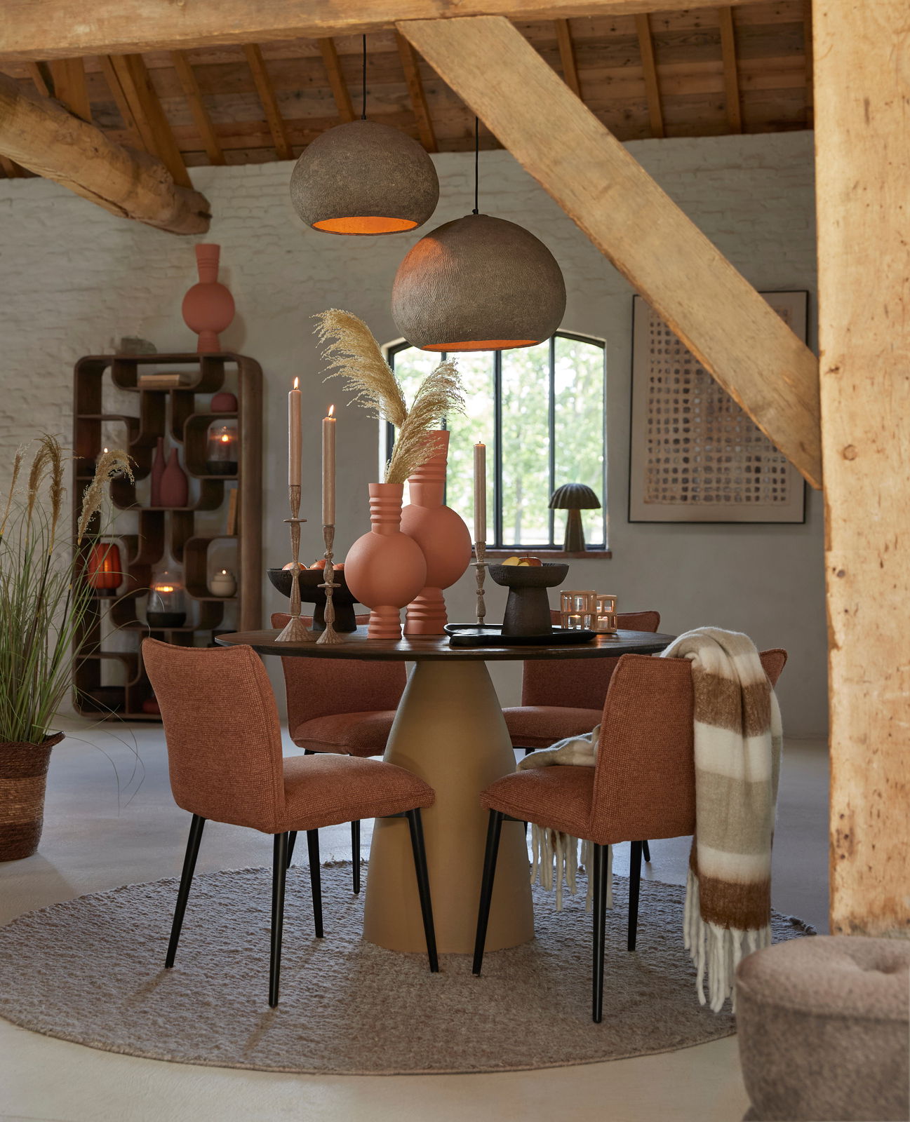 Moderner Esstisch mit runder Tischplatte und konischem Sockel, umgeben von vier gepolsterten Stühlen in einem gemütlichen Raum mit rustikalen Holzbalken, aus der Frontalperspektive.