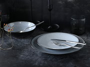 Elegantes Tellerset in dunkler, stilvoller Umgebung, bestehend aus einem großen und einem kleinen Teller mit feiner Musterung, dazu passendes Besteck und eine Schale, seitliche Perspektive.