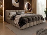 Een gestoffeerd bed in een stijlvolle slaapkamer, gezien vanaf de zijkant, met een beige hoofdeinde en een doorgestikt bedframe, versierd met kussens en dekens.