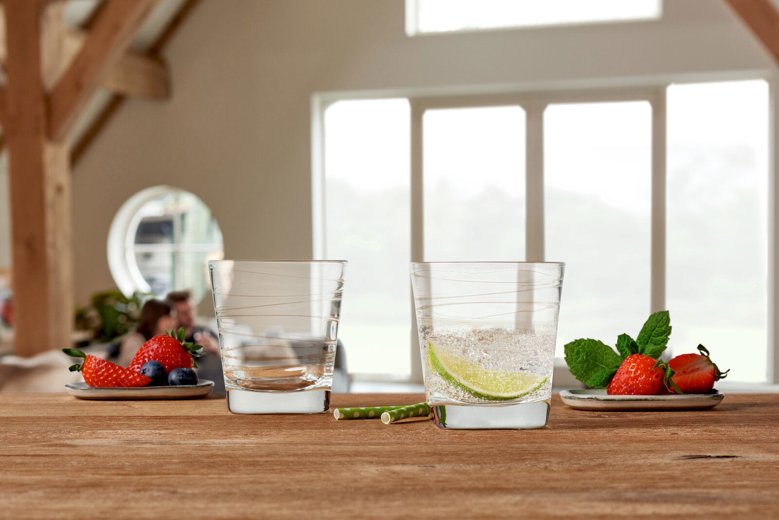 Zwei Trinkgläser Vario 250ml auf einem Holztisch, gefüllt mit Wasser und Limettenscheiben, umgeben von frischen Erdbeeren und Minzblättern, in einem hellen Raum mit großen Fenstern, aus der Vorderansicht fotografiert.