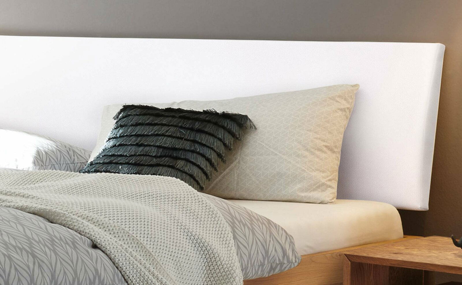 Nahaufnahme eines Bettgestells mit weißem Kopfteil, grauem Kissen mit Fransen und beigem Kissen, aus der Seitenperspektive fotografiert.