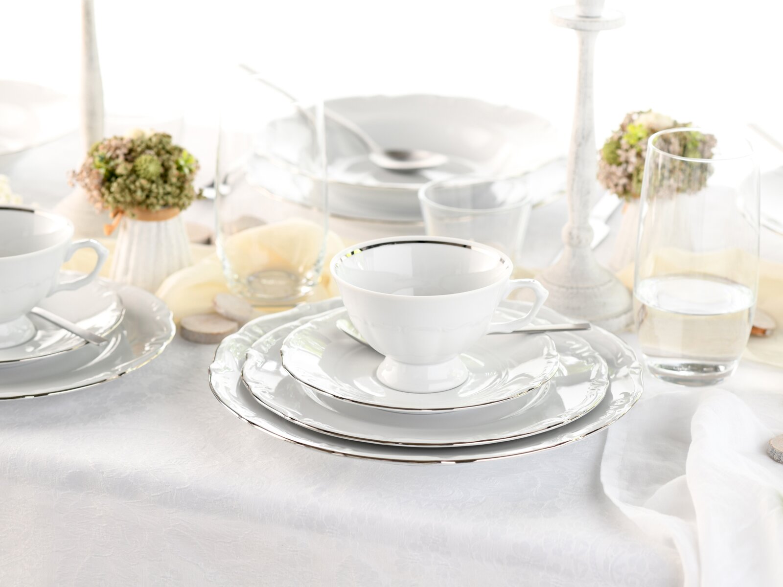 Elegantes weißes Porzellan-Kaffeegedeck mit Tasse und Untertasse auf gestapelten Tellern mit silbernem Rand, gedeckter Tisch mit Gläsern und Kerzenhalter im Hintergrund, Perspektive: leicht von vorne in Augenhöhe