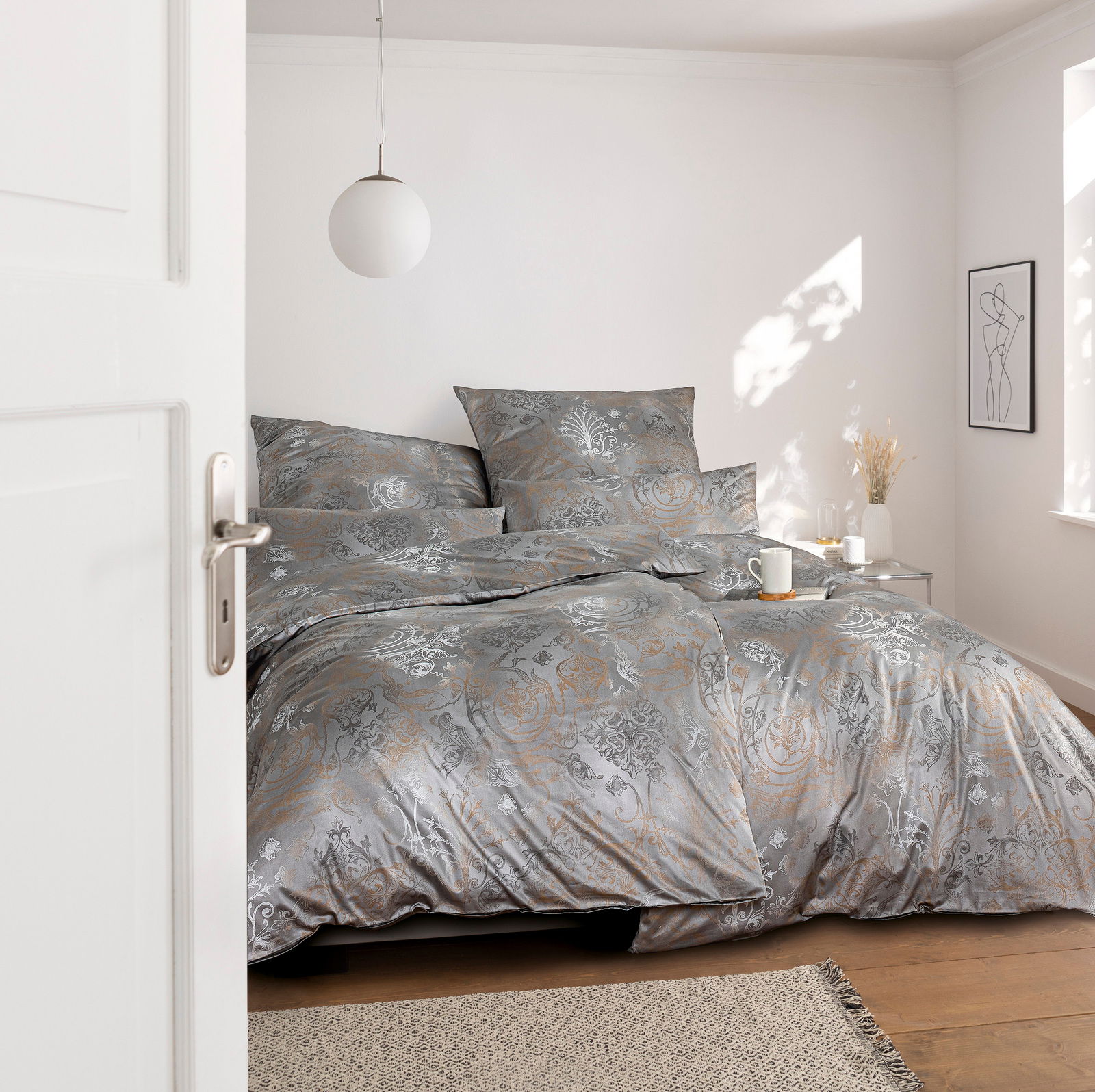 Blick in ein stilvolles Schlafzimmer mit eleganter Bettwäsche in Grau- und Goldtönen, dekoriert mit floralen Mustern. Die Bettwäsche ist auf einem Doppelbett arrangiert, umgeben von minimalistischer Dekoration und natürlichem Licht.