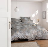 Blick in ein stilvolles Schlafzimmer mit eleganter Bettwäsche in Grau- und Goldtönen, dekoriert mit floralen Mustern. Die Bettwäsche ist auf einem Doppelbett arrangiert, umgeben von minimalistischer Dekoration und natürlichem Licht.