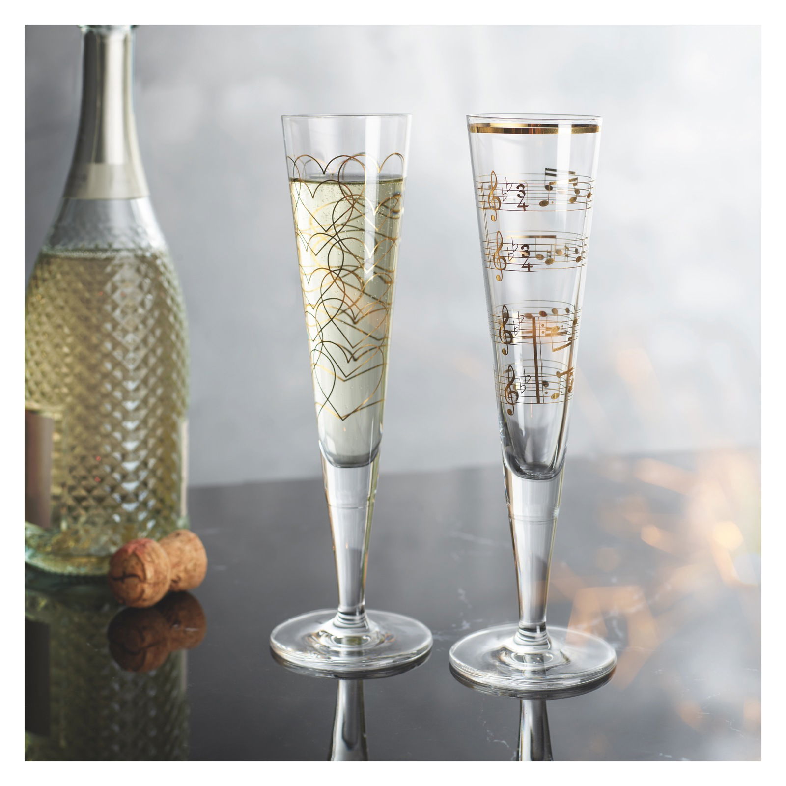 Zwei elegante Champagnergläser mit goldenen Verzierungen, eines mit Herzmustern und das andere mit Notenlinien, auf einem Tisch mit einer Flasche Sekt im Hintergrund. Frontale Perspektive.