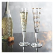 Twee elegante champagneglazen met gouden versieringen, het ene met hartjesmotieven en het andere met notenbalken, op een tafel met op de achtergrond een fles mousserende wijn. Vooraanzicht.