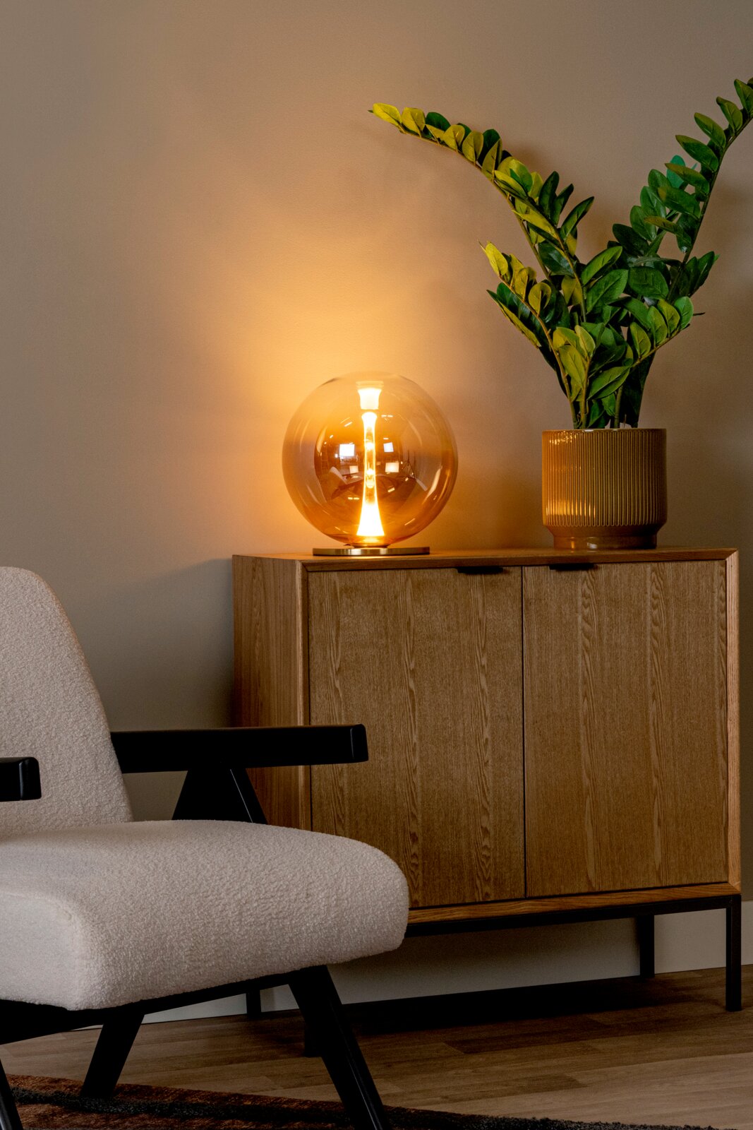 Leuchtende Kugel-Tischlampe aus amberfarbenem Glas auf Holz-Sideboard neben grünem Blumentopf, frontale Perspektive auf das Möbel im Wohnzimmer