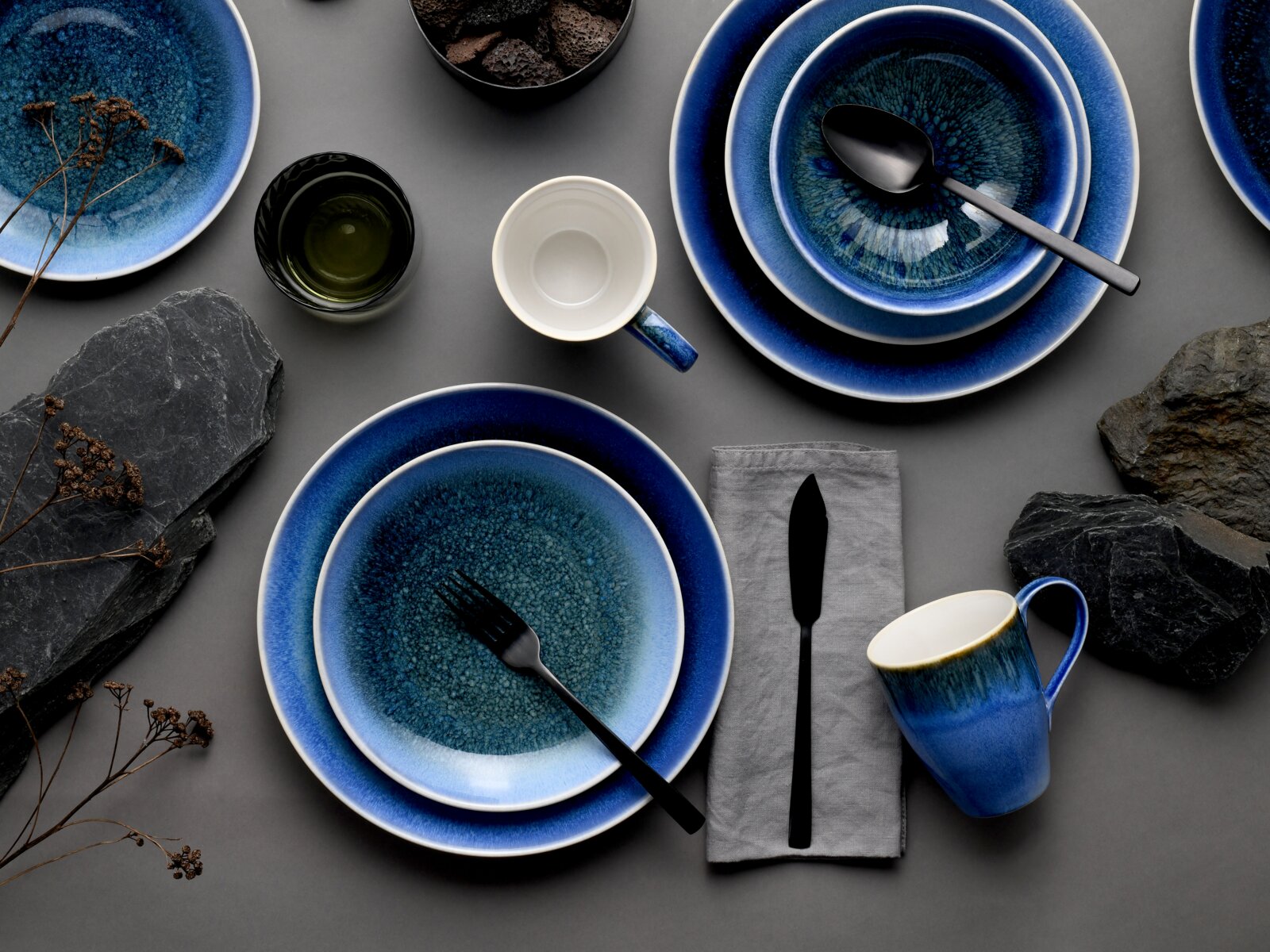 Draufsicht auf ein blau glasiertes Keramik-Geschirrset mit Tellern, Schalen und Tassen, arrangiert mit schwarzem Besteck auf grauem Hintergrund.