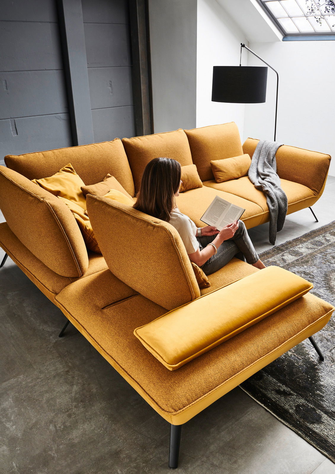 Eckgarnitur in senfgelber Farbe mit mehreren Kissen, auf der eine Person sitzt und ein Buch liest. Perspektive von oben.