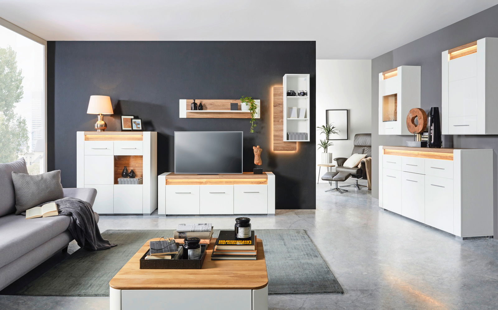 Modernes Wohnzimmer mit TV-Unterteil in Weiß und Holzoptik, frontal betrachtet. Das TV-Unterteil steht vor einer dunklen Wand und ist von weiteren Möbelstücken umgeben.