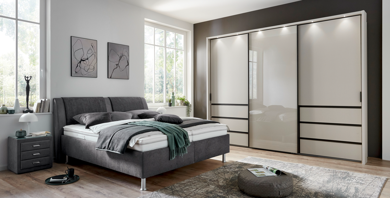 Modernes Polsterbett in einem stilvollen Schlafzimmer, seitliche Perspektive mit grauem Kopfteil und passendem Nachttisch.