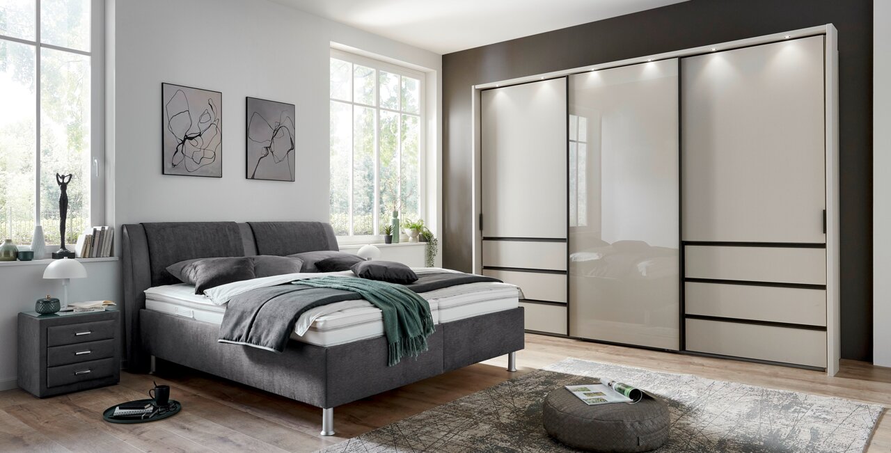 Modernes Polsterbett in einem stilvollen Schlafzimmer, seitliche Perspektive mit grauem Kopfteil und passendem Nachttisch.