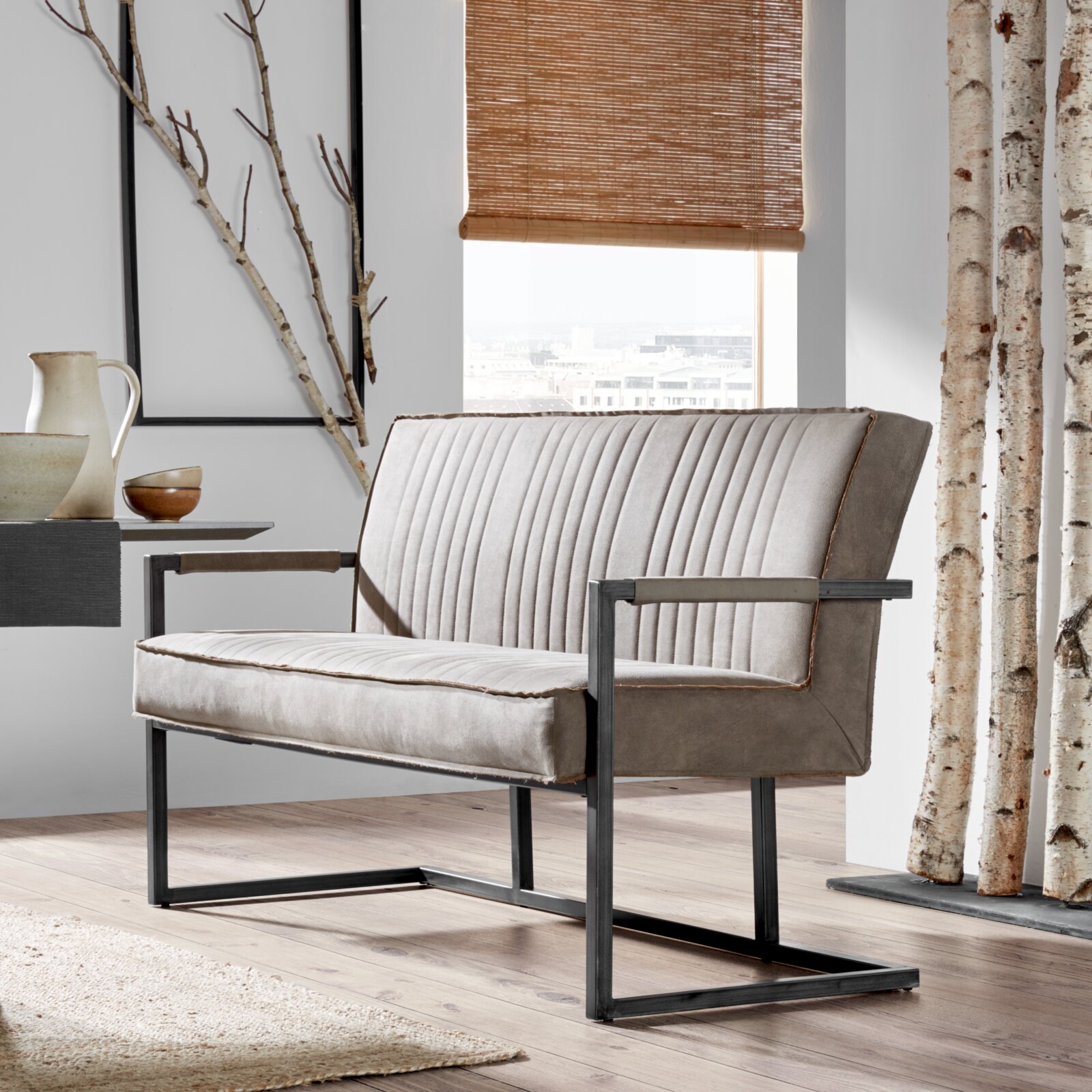 Moderne grijze bank met metalen frame, gezien vanaf de zijkant, in een stijlvol ingerichte ruimte met een houten vloer en decoratieve elementen.