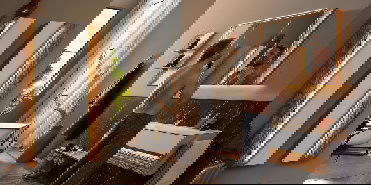 Das Bild zeigt ein modernes Badezimmer. Ein Mann steht vor einem großen Spiegel und rasiert sich. Der Waschtisch darunter hat eine Holzoptik und weiße Fronten. Rechts und links an der Wand hängen schmale Schränke mit Beleuchtung. Im Hintergrund ist eine B