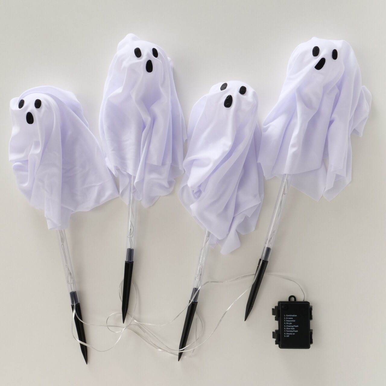 Vier weiße Halloween-Geisterfiguren aus Stoff mit schwarzen Augen und Mund auf transparenten Stäben, verbunden mit Kabeln und einem schwarzen Batteriekasten, Draufsicht auf hellem Hintergrund