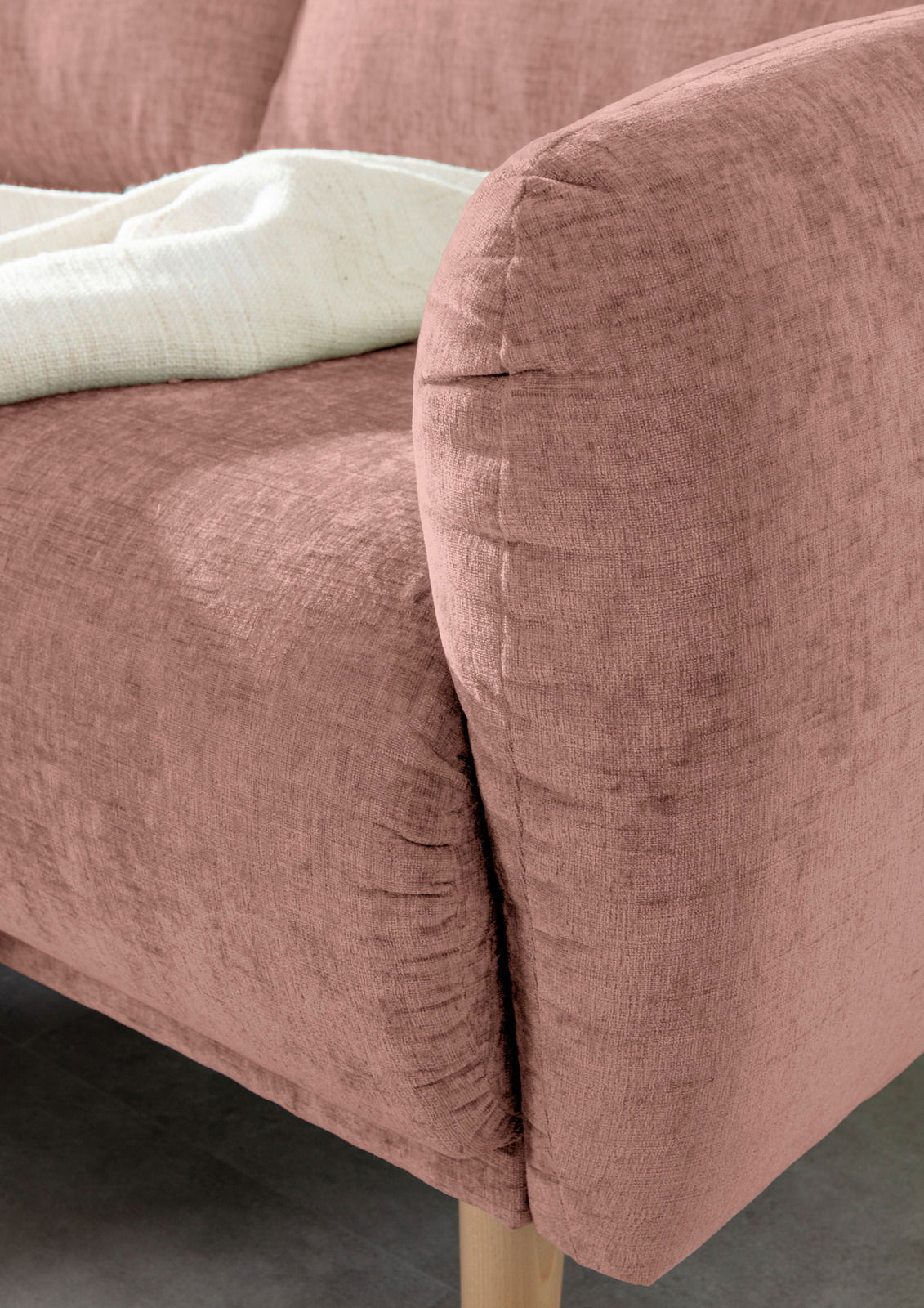 Nahaufnahme einer rosafarbenen Sofaecke mit weicher Textur und Holzbeinen, von der Seite betrachtet.