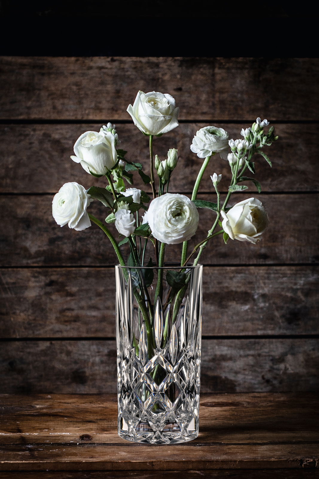 Kristallvase mit weißen Blumen, frontal auf einem Holztisch vor Holzwand