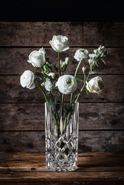 Kristallvase mit weißen Blumen, frontal auf einem Holztisch vor Holzwand