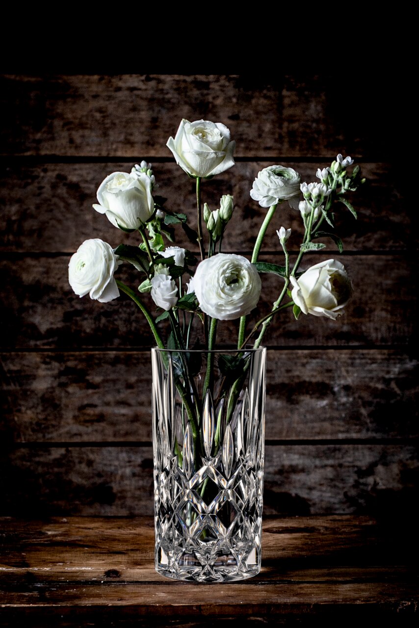 Kristallvase mit weißen Blumen, frontal auf einem Holztisch vor Holzwand