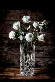 Kristallvase mit weißen Blumen, frontal auf einem Holztisch vor Holzwand