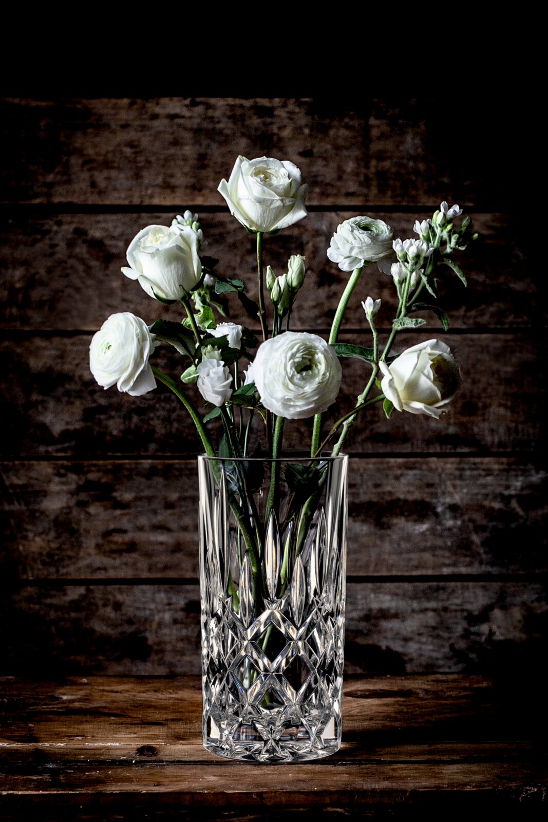 Kristallen vaas met witte bloemen, vooraanzicht op een houten tafel voor een houten wand