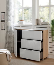 Moderne Kommode mit drei weißen Hochglanz-Schubladen und einer offenen Tür, aus hellem Holz, in einem hellen Raum mit großen Fenstern, seitliche Perspektive.
