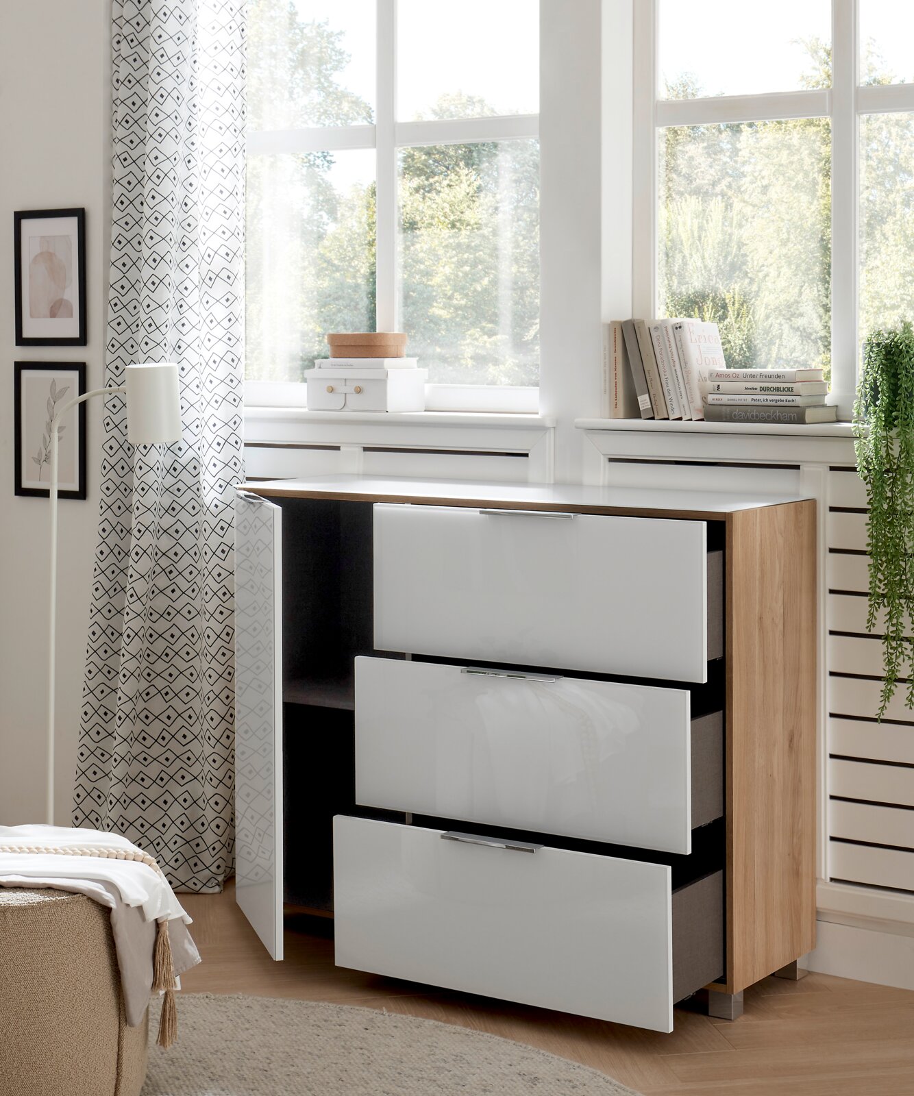 Moderne Kommode mit drei weißen Hochglanz-Schubladen und einer offenen Tür, aus hellem Holz, in einem hellen Raum mit großen Fenstern, seitliche Perspektive.