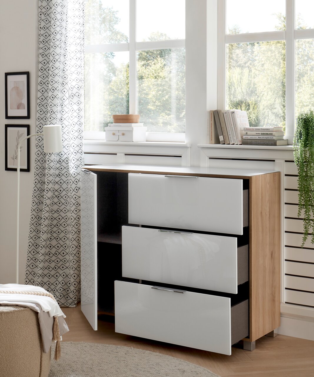 Moderne Kommode mit drei weißen Hochglanz-Schubladen und einer offenen Tür, aus hellem Holz, in einem hellen Raum mit großen Fenstern, seitliche Perspektive.