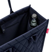 Nahaufnahme einer dunkelblauen Einkaufstasche mit Rautenmuster, die eine Flasche in einem Seitenfach zeigt.