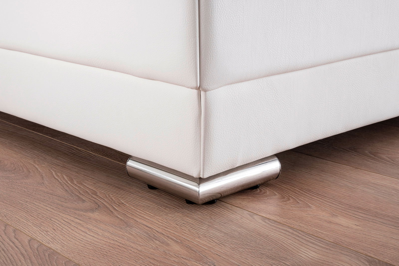Nahaufnahme der Ecke eines weißen Polsterbetts mit metallischen Füßen auf einem Holzboden, Perspektive von unten.
