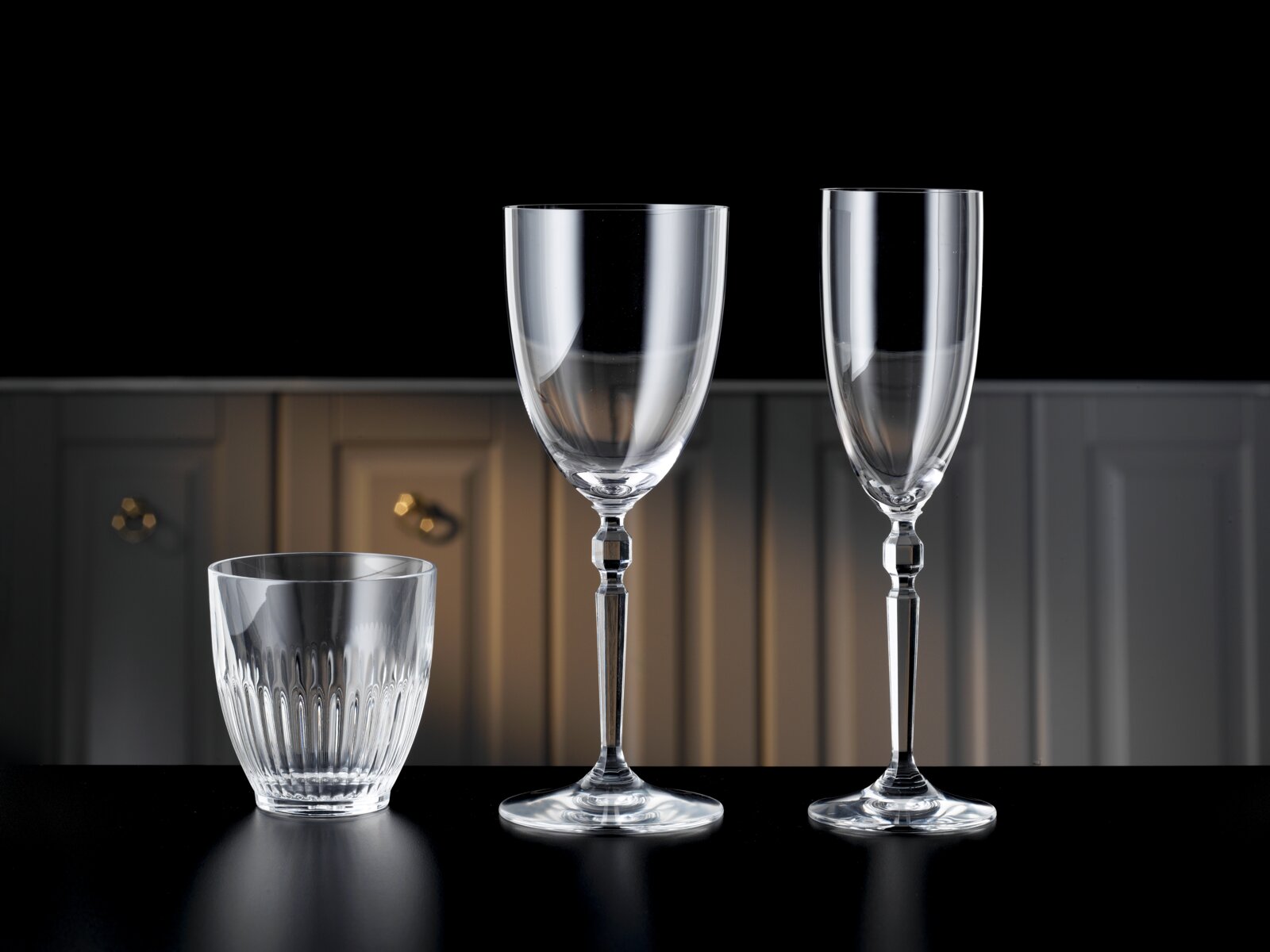 Drei transparente Trinkgläser (zwei hohe Stielgläser und ein geriffeltes Tumblerglas) auf schwarzer Oberfläche vor dunklem Hintergrund, frontal in leicht niedriger Perspektive aufgenommen.