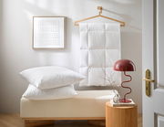 Ein stilvoll eingerichtetes Zimmer mit einem weißen 3-Kammer Kopfkissen und einer Daunendecke, die auf einem Kleiderbügel hängt. Im Vordergrund steht eine rote Lampe auf einem kleinen Tisch. Perspektive: seitlich.