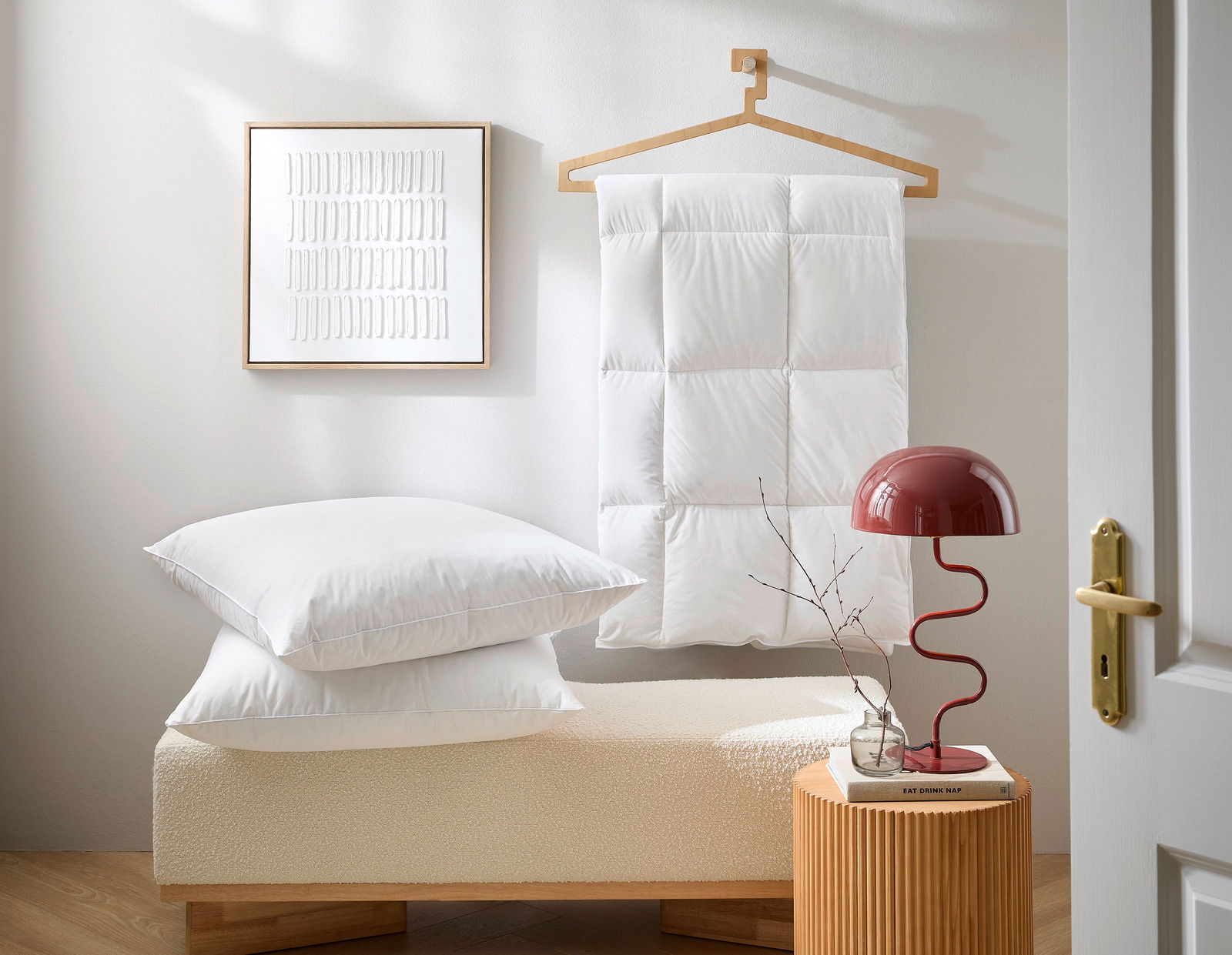 billerbeck 3-Kammer Kopfkissen Daune Polarstern Ein stilvoll eingerichtetes Zimmer mit einem weißen 3-Kammer Kopfkissen und einer Daunendecke, die auf einem Kleiderbügel hängt. Im Vordergrund steht eine rote Lampe auf einem kleinen Tisch. Perspektive: seitlich.