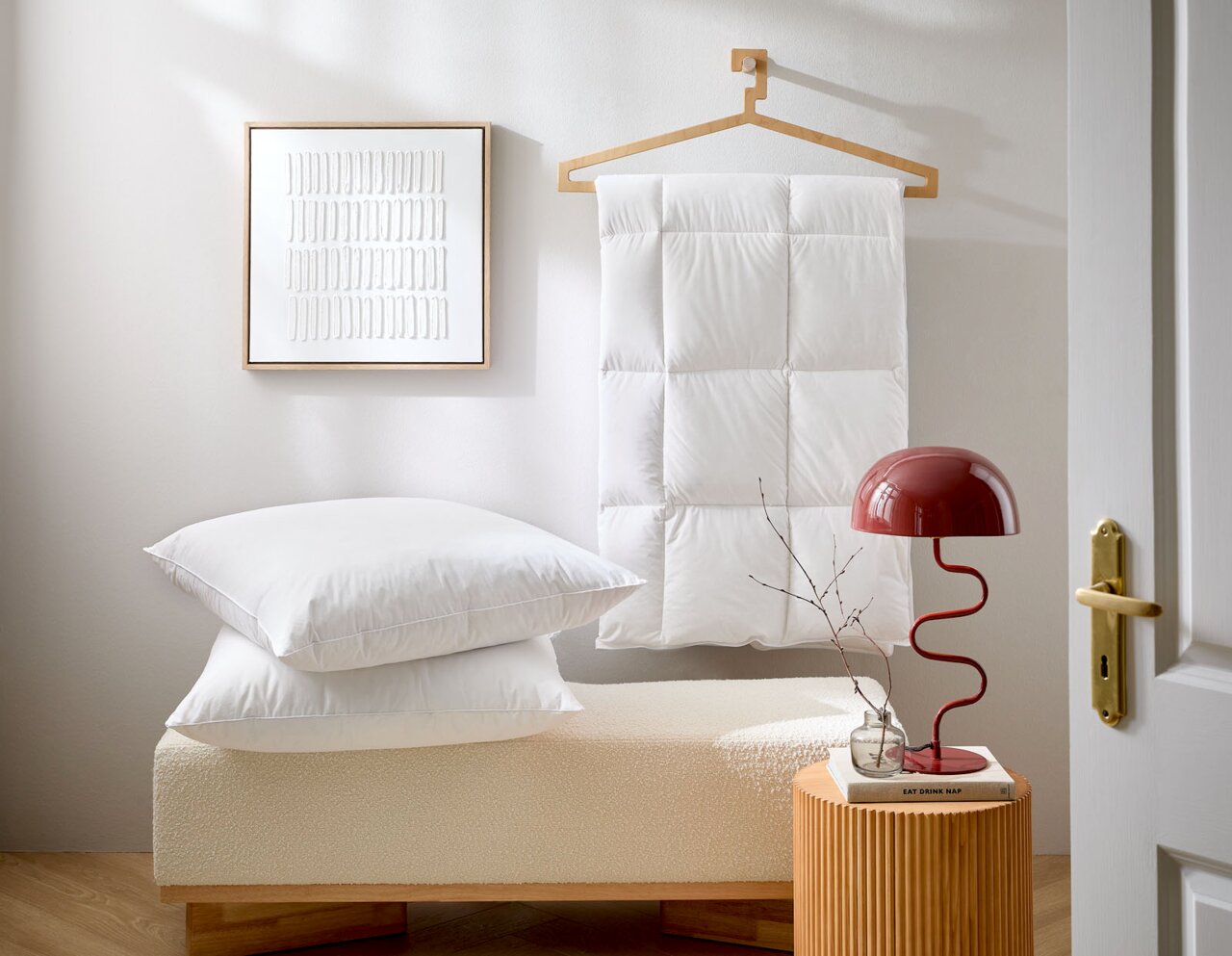 Ein stilvoll eingerichtetes Zimmer mit einem weißen 3-Kammer Kopfkissen und einer Daunendecke, die auf einem Kleiderbügel hängt. Im Vordergrund steht eine rote Lampe auf einem kleinen Tisch. Perspektive: seitlich.